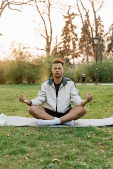 Young man in lotus pose meditating outdoors on a yoga mat at sunset, promoting relaxation and wellness.