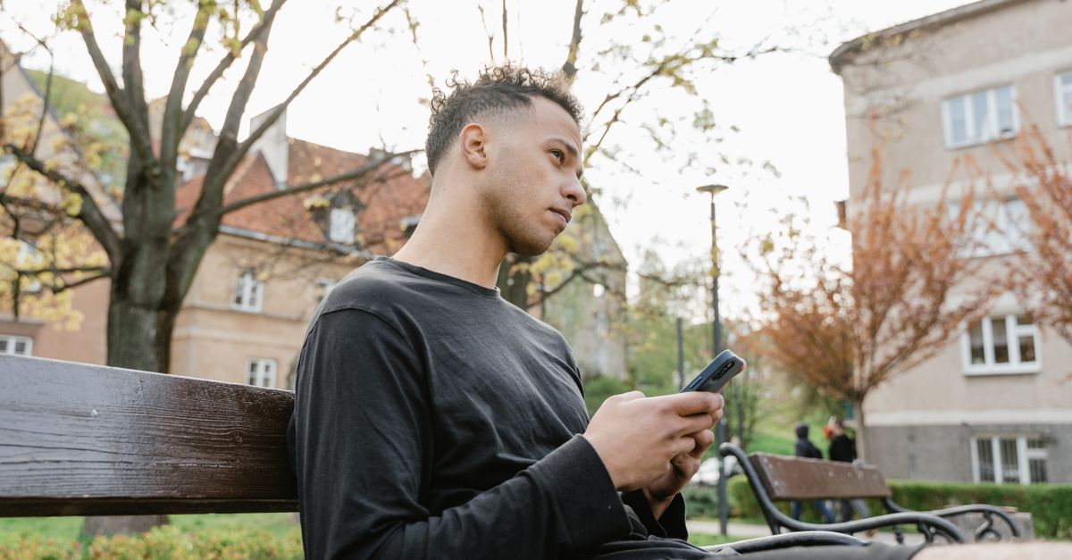 A young Ghanaian man reading a Bible on a smartphone while sitting on a bench in a park in Ho, with a church building visible in the background