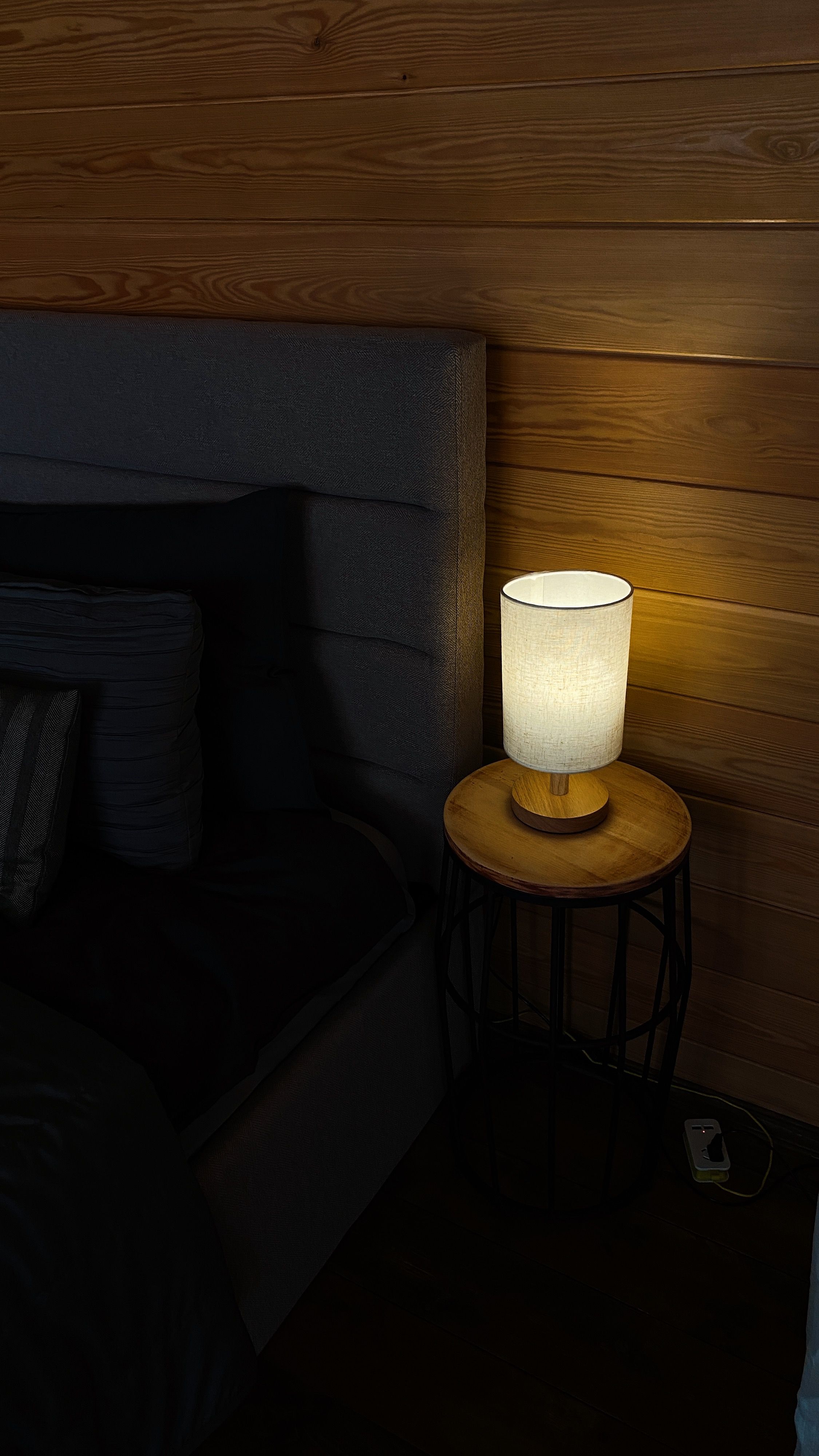 Glowing shaded lamp on a wooden bedside table against a dark paneled wall