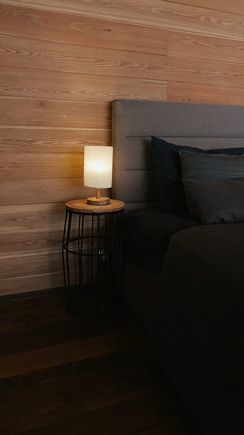 Bed with cushion placed near wooden wall and glowing lamp on bedside table