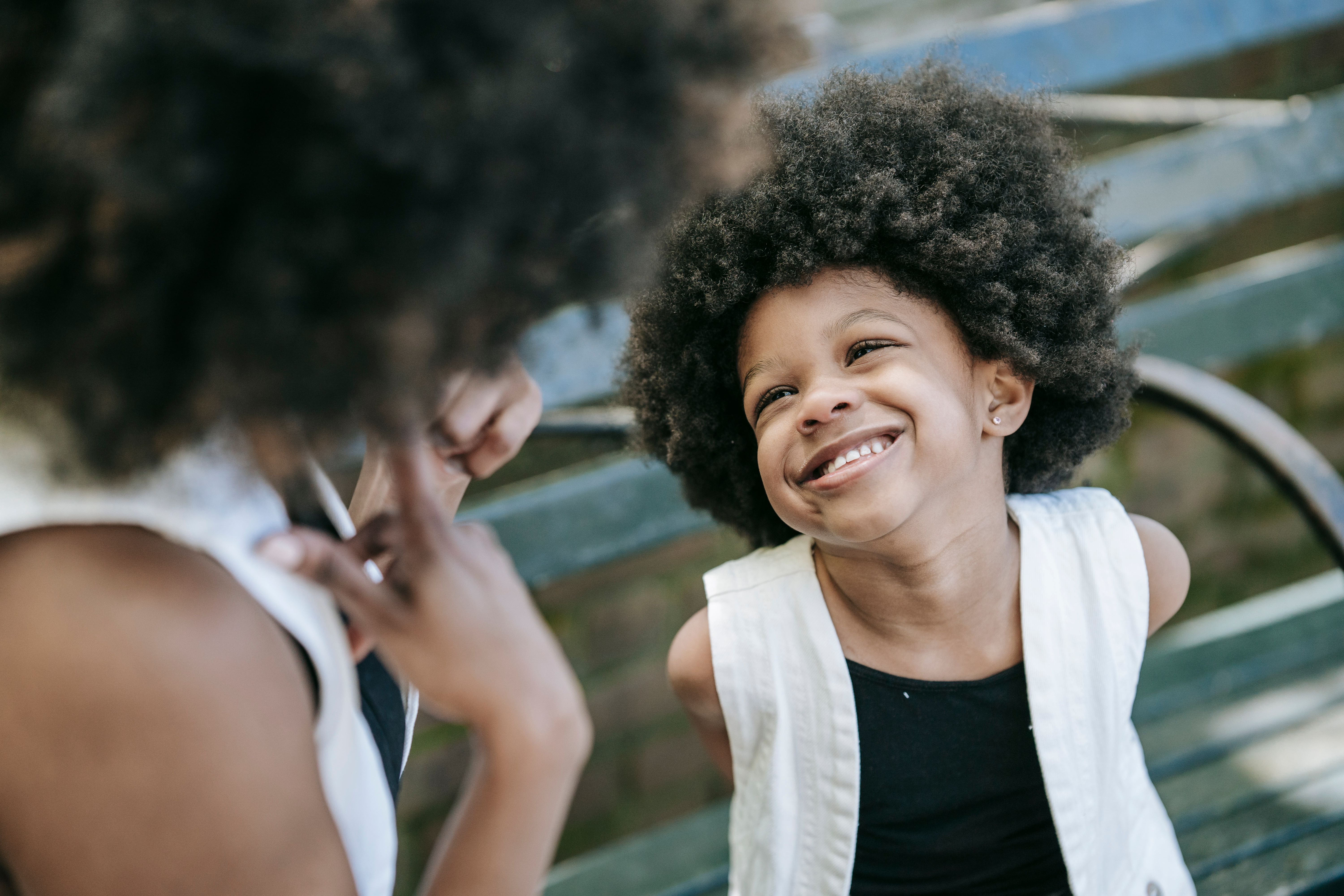 A cheerful child with afro hair joyfully smiles while interacting outside on a bench.