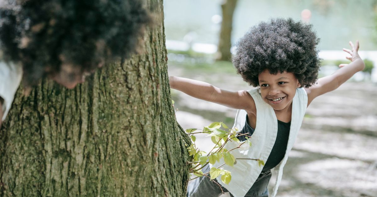 An African American girl playing hide and seek around a tree trunk in a sunny park.