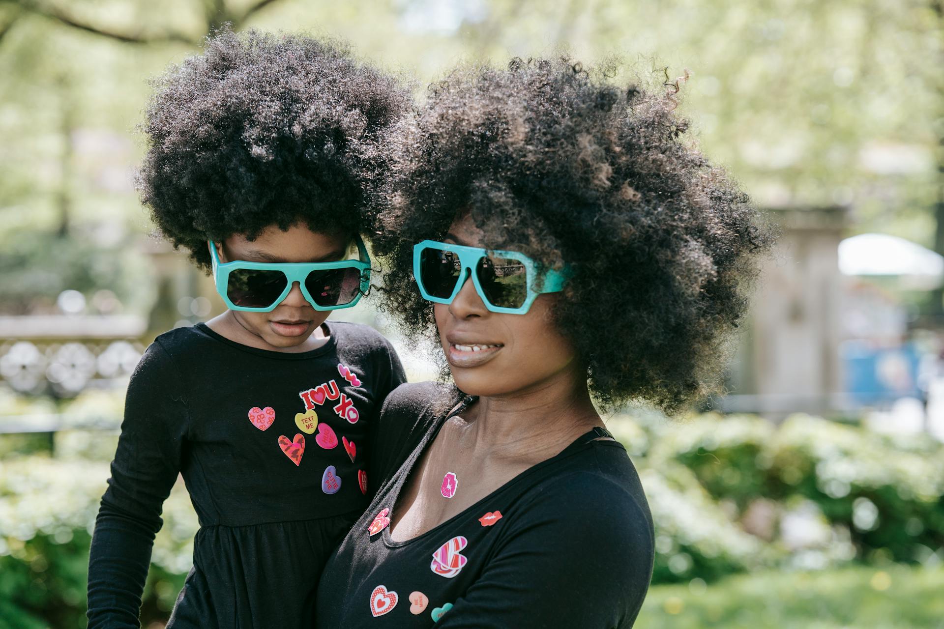 A mother and daughter with matching afro hairstyles and sunglasses share a joyful moment outdoors.