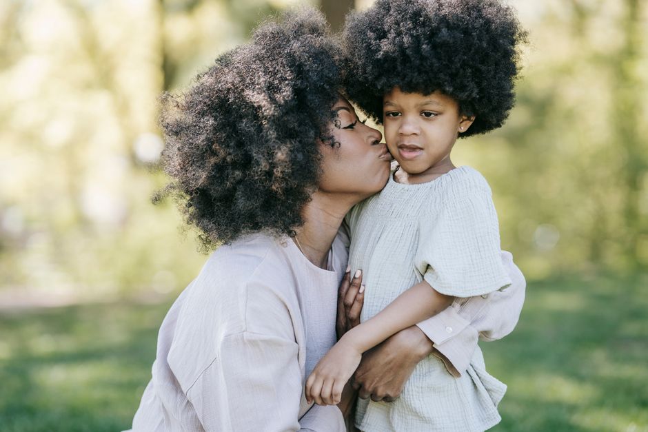 Tender moment of a mother kissing her daughter in a sunny park setting.