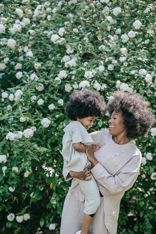 A Woman Carrying Her Daughter while Standing Near the White Flowers with Green Leaves