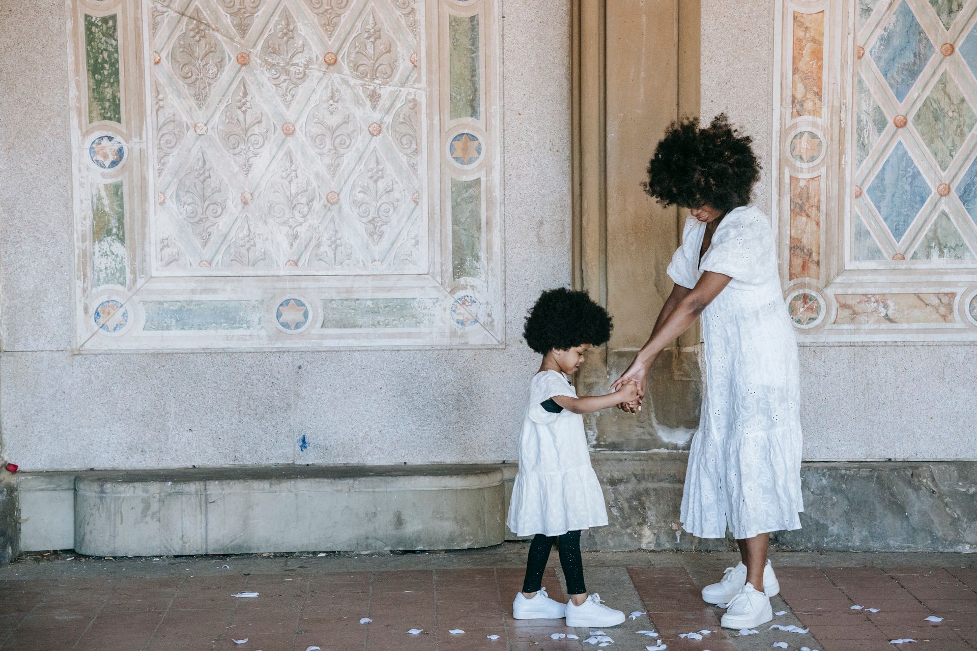 A stylish mother and daughter in matching outfits share a moment, holding hands near a decorated wall.