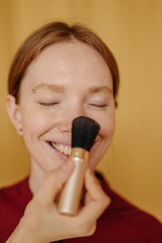 Close-up of a smiling woman having makeup applied with a brush indoors.