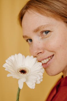 Close-up portrait of a smiling woman holding a white gerbera daisy against a yellow background.