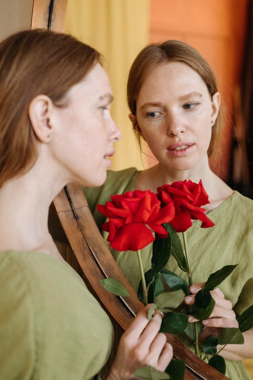 Close Up Photo of Woman Holding Red Flower in Front of a Mirror