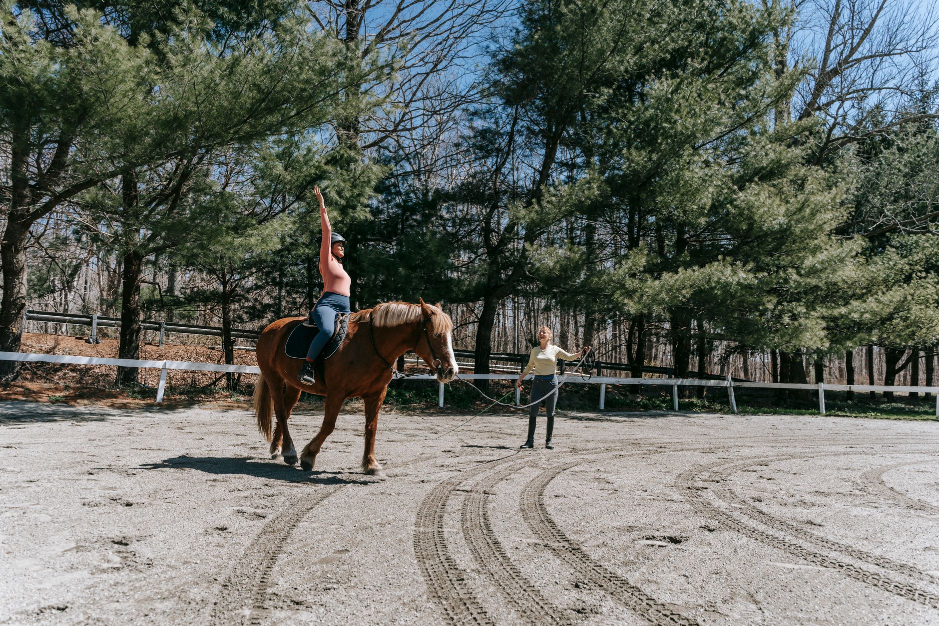 A woman practices horseback riding under the guidance of an instructor in a forested outdoor arena.