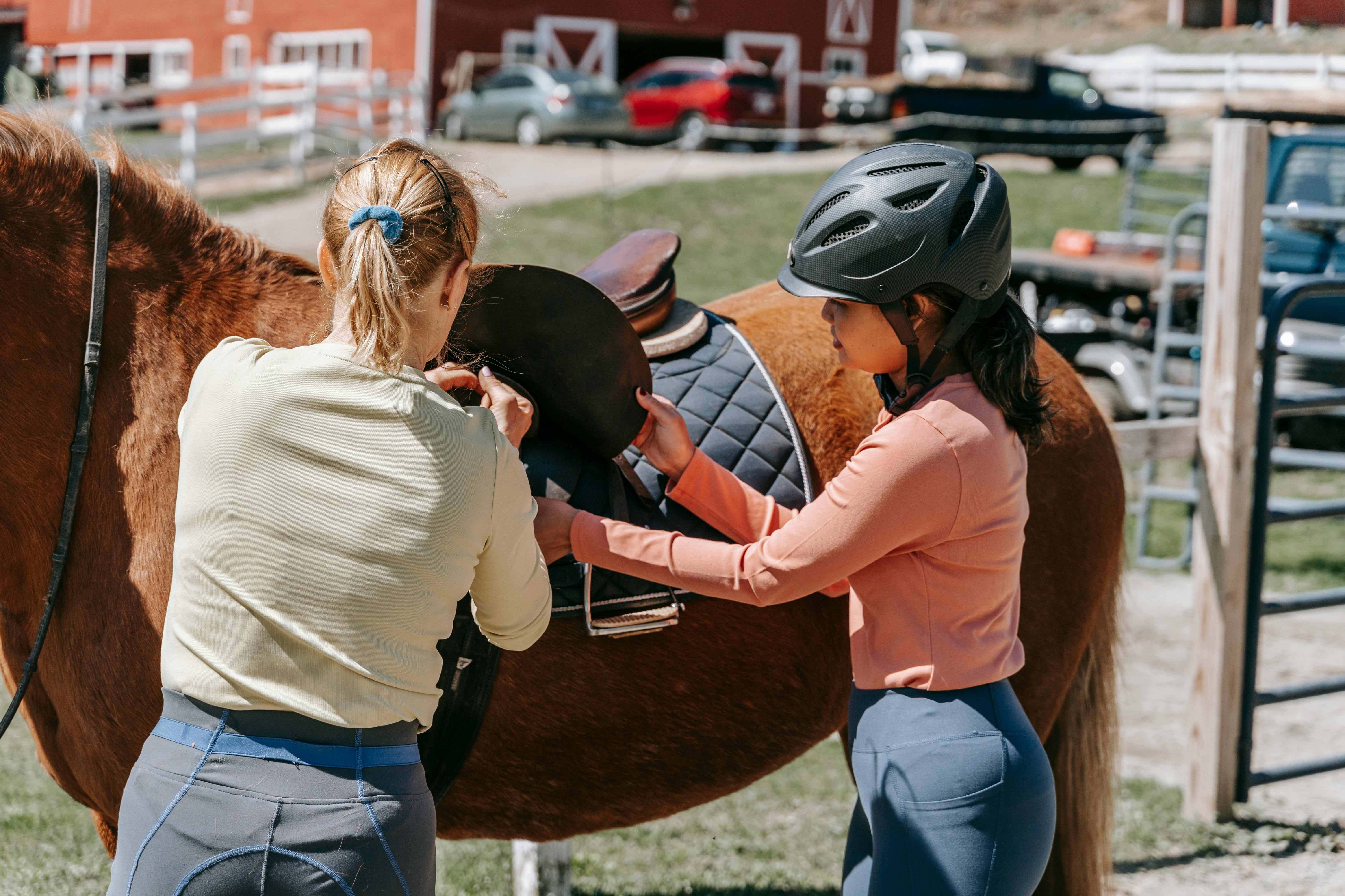 Two women preparing a horse for riding on a sunny day at a farm.