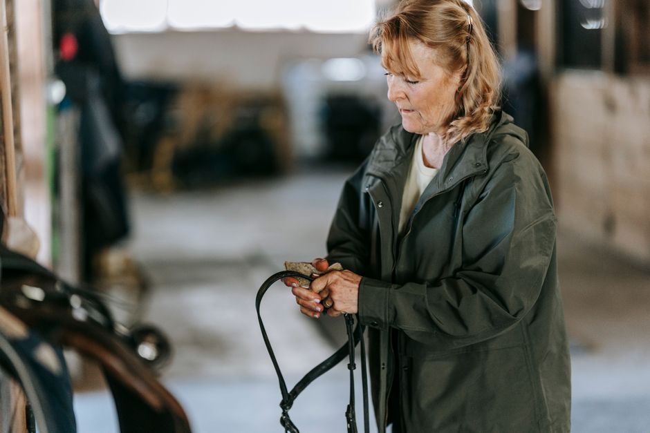 Elderly woman organizing horse tack in a stable, wearing a green jacket, focused on handling reins.