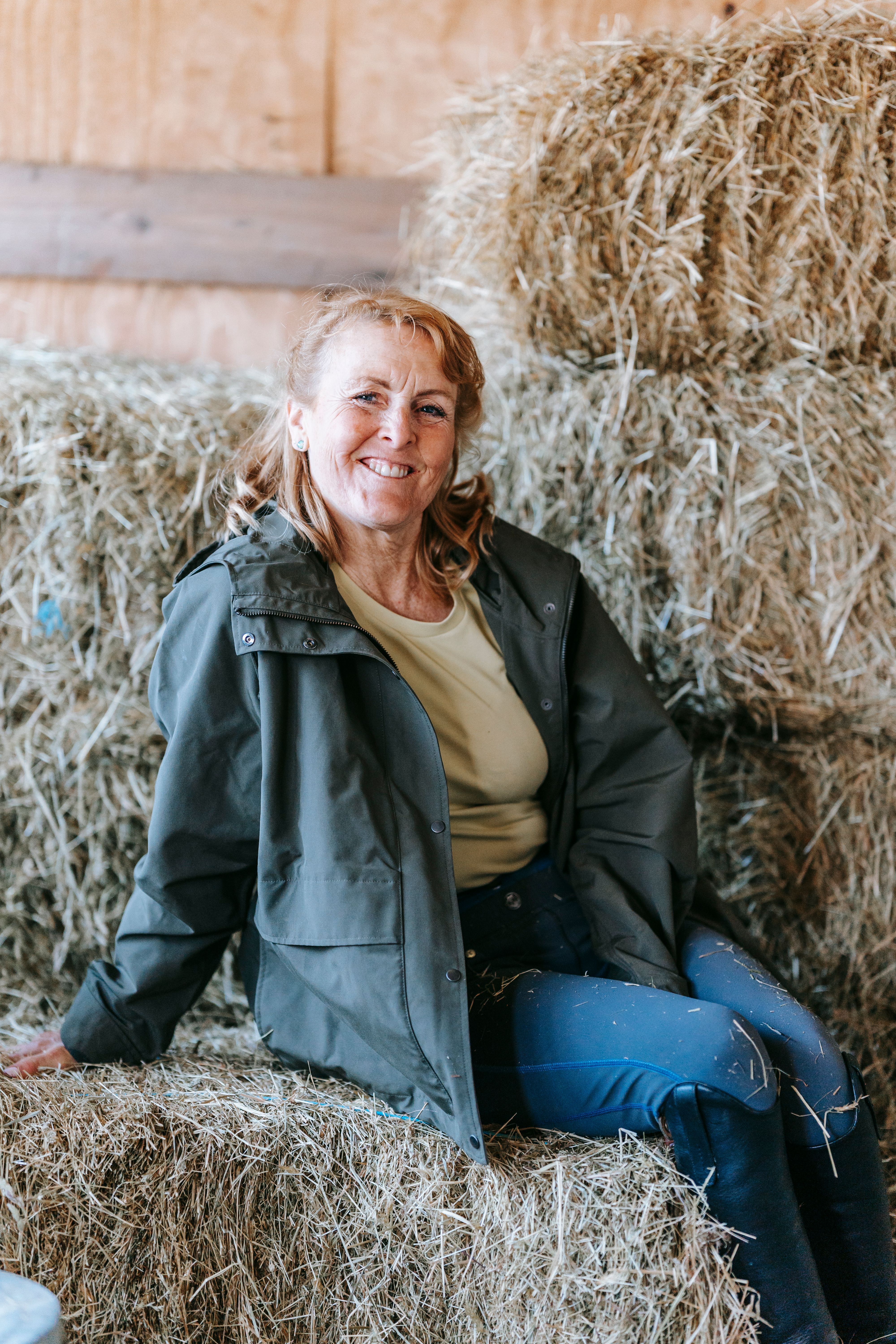 Smiling woman in an outdoor jacket seated on hay bales, exuding warmth and rustic charm.