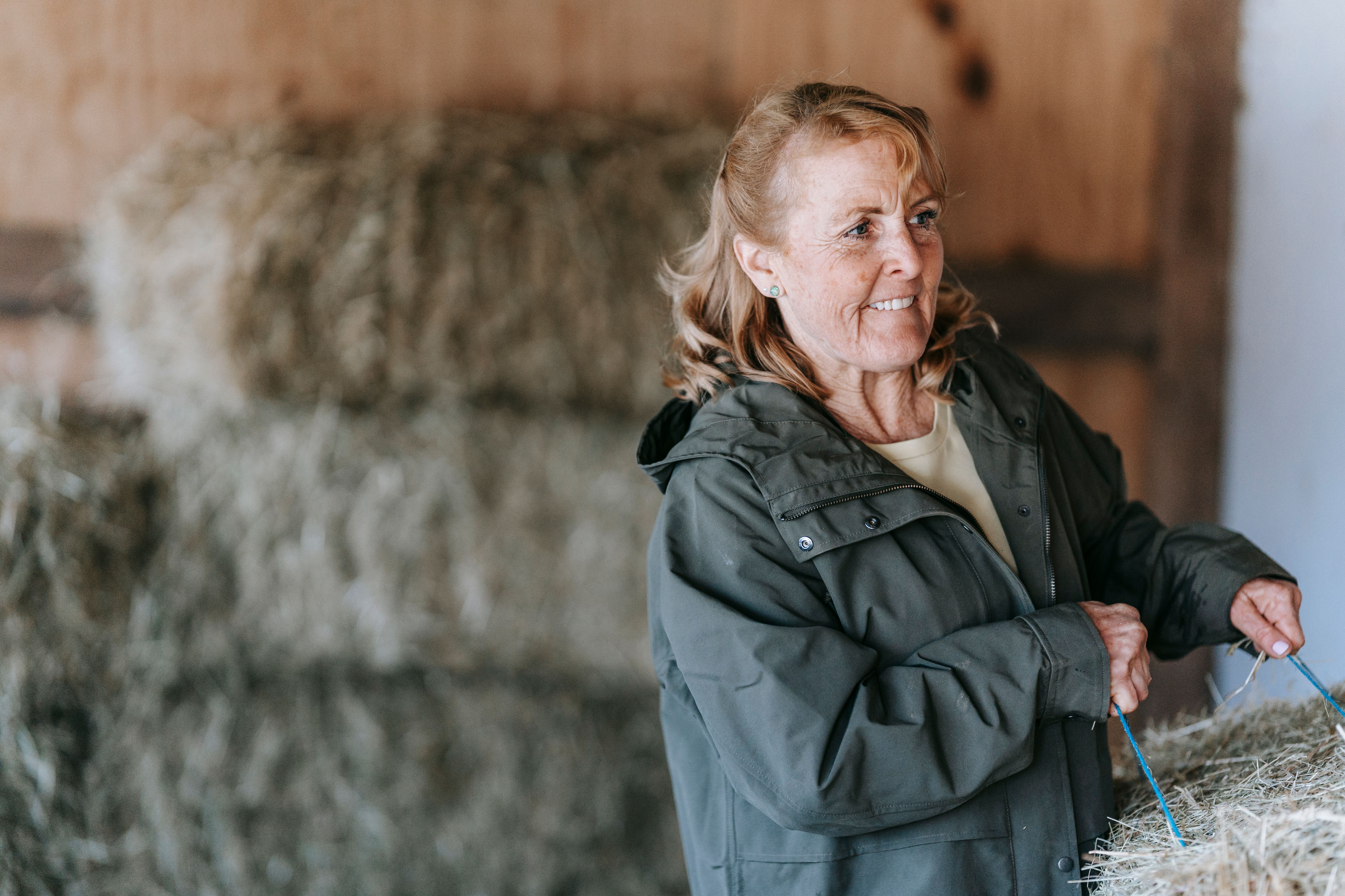 Elderly woman in a barn handling hay, showcasing rural lifestyle.