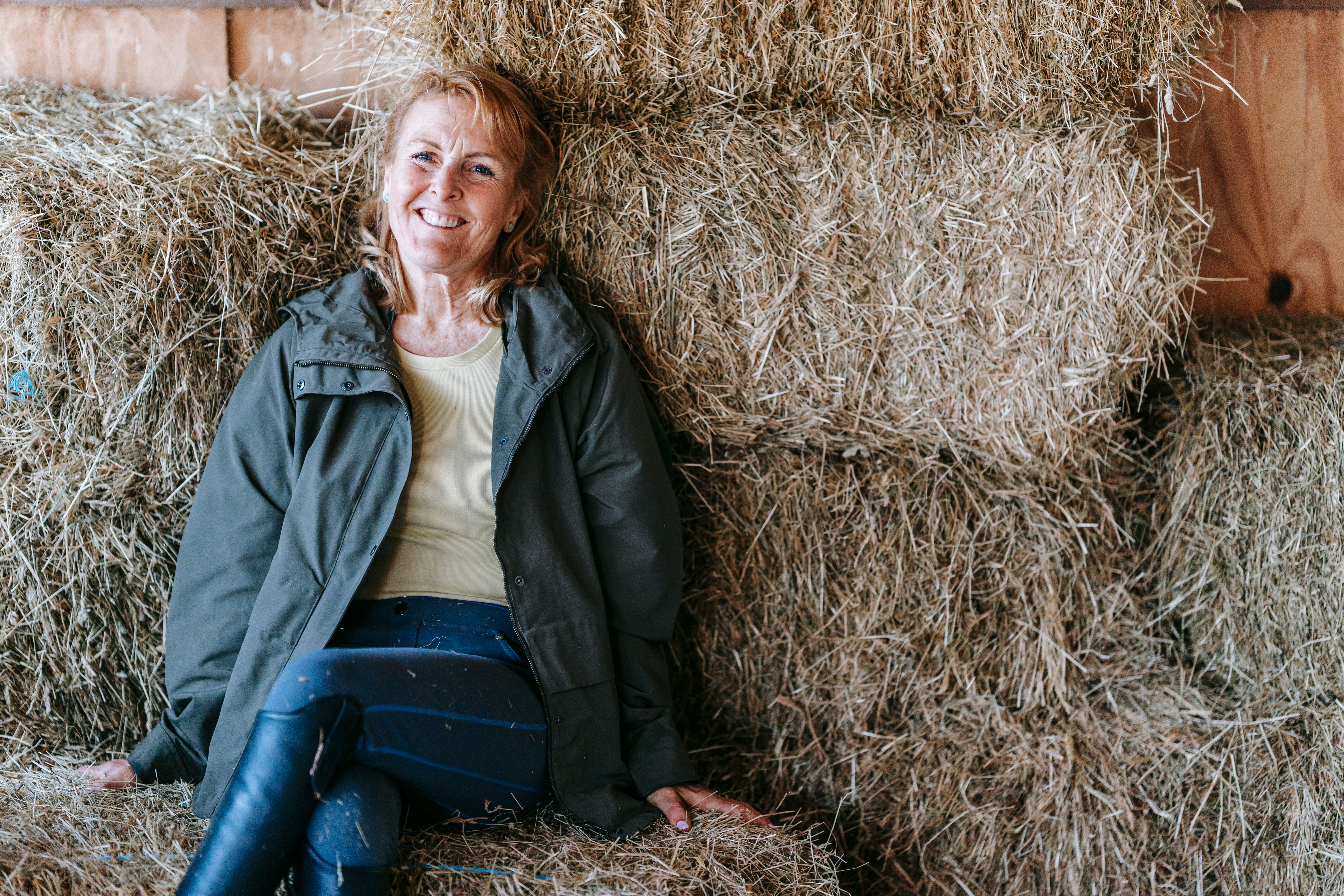 Senior woman with a warm smile sitting on hay bales in a barn.