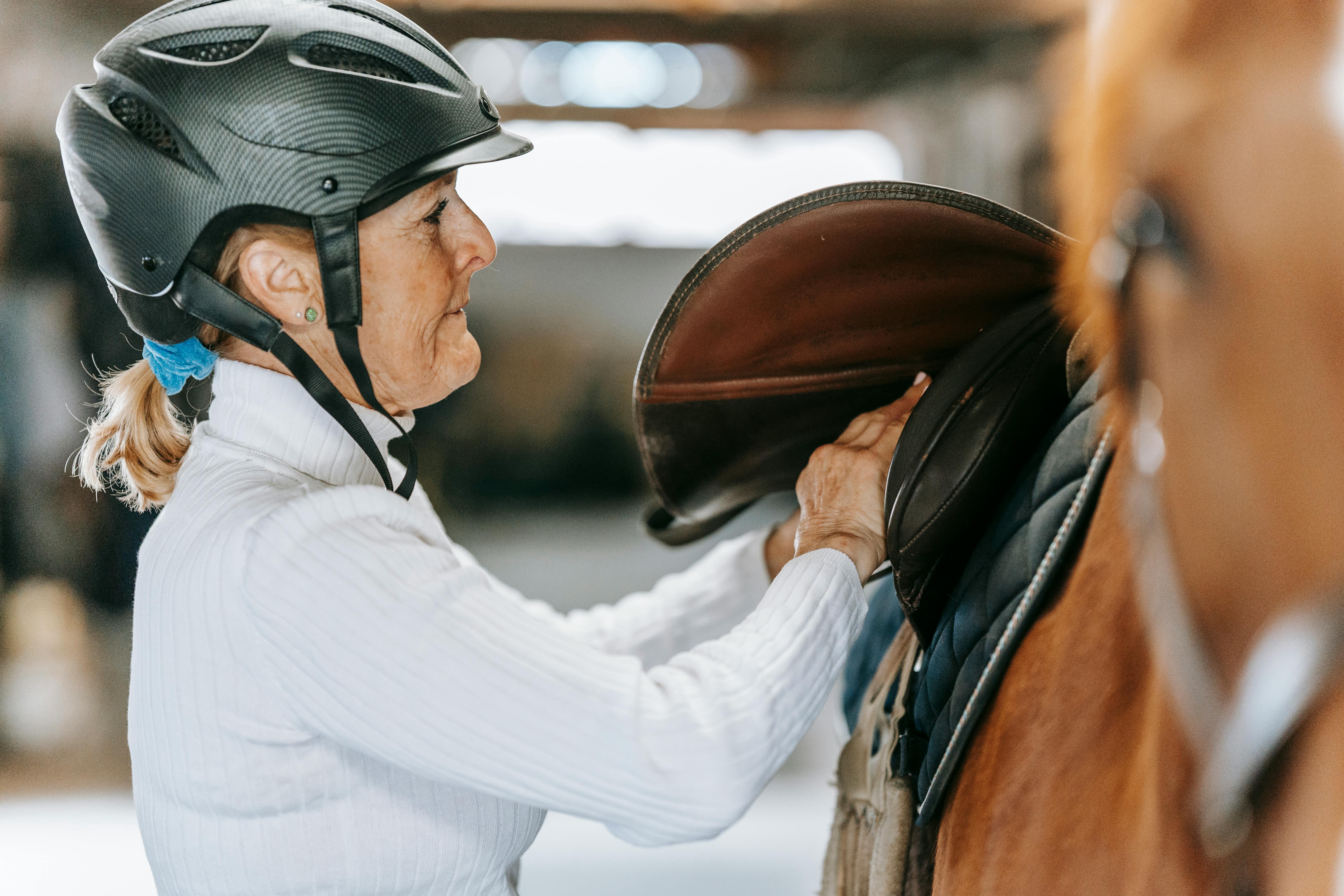 Adult woman prepares horse for riding by adjusting saddle in stable.