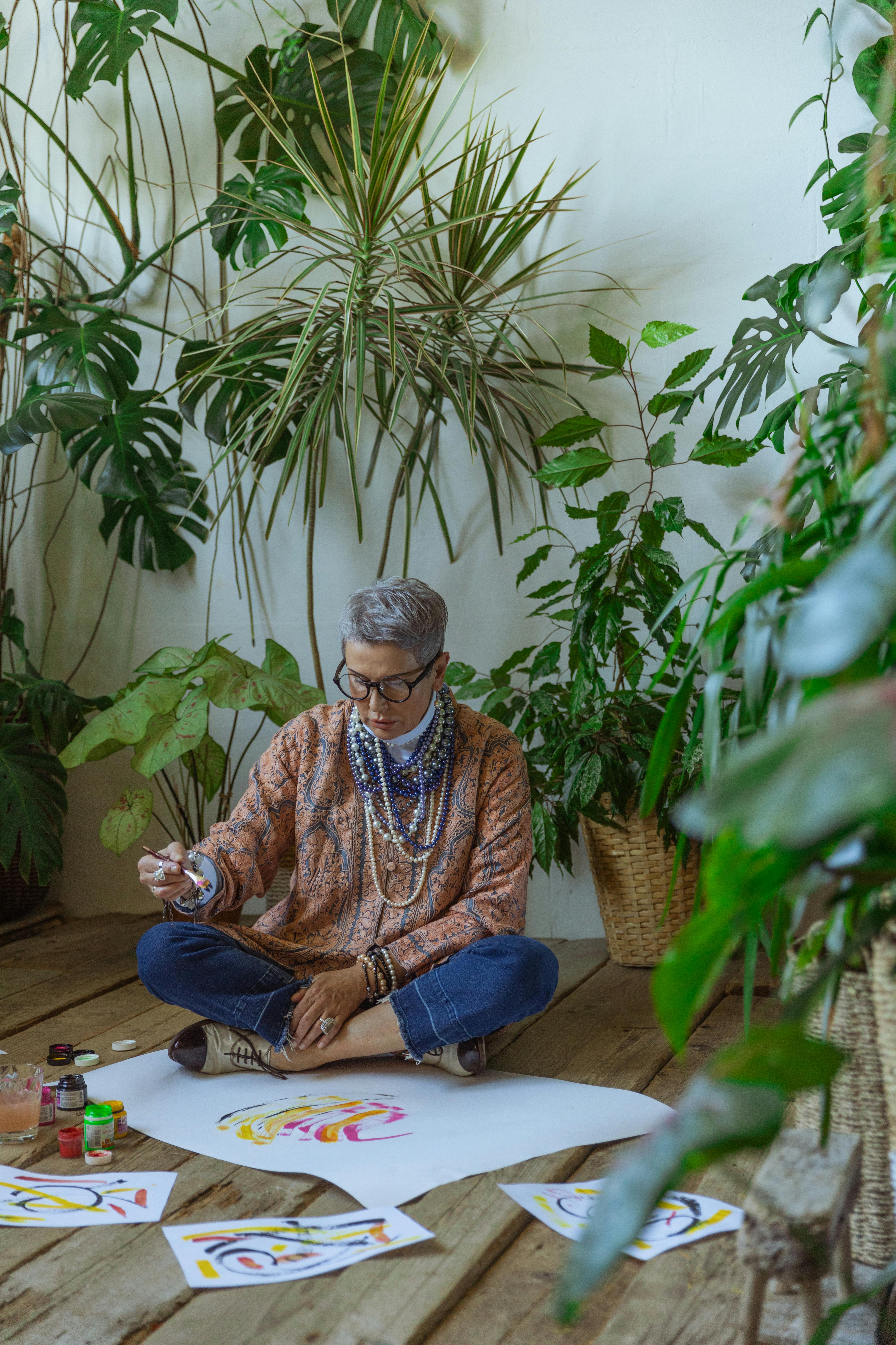 Senior woman painting surrounded by lush indoor plants, showcasing creativity in a natural setting.