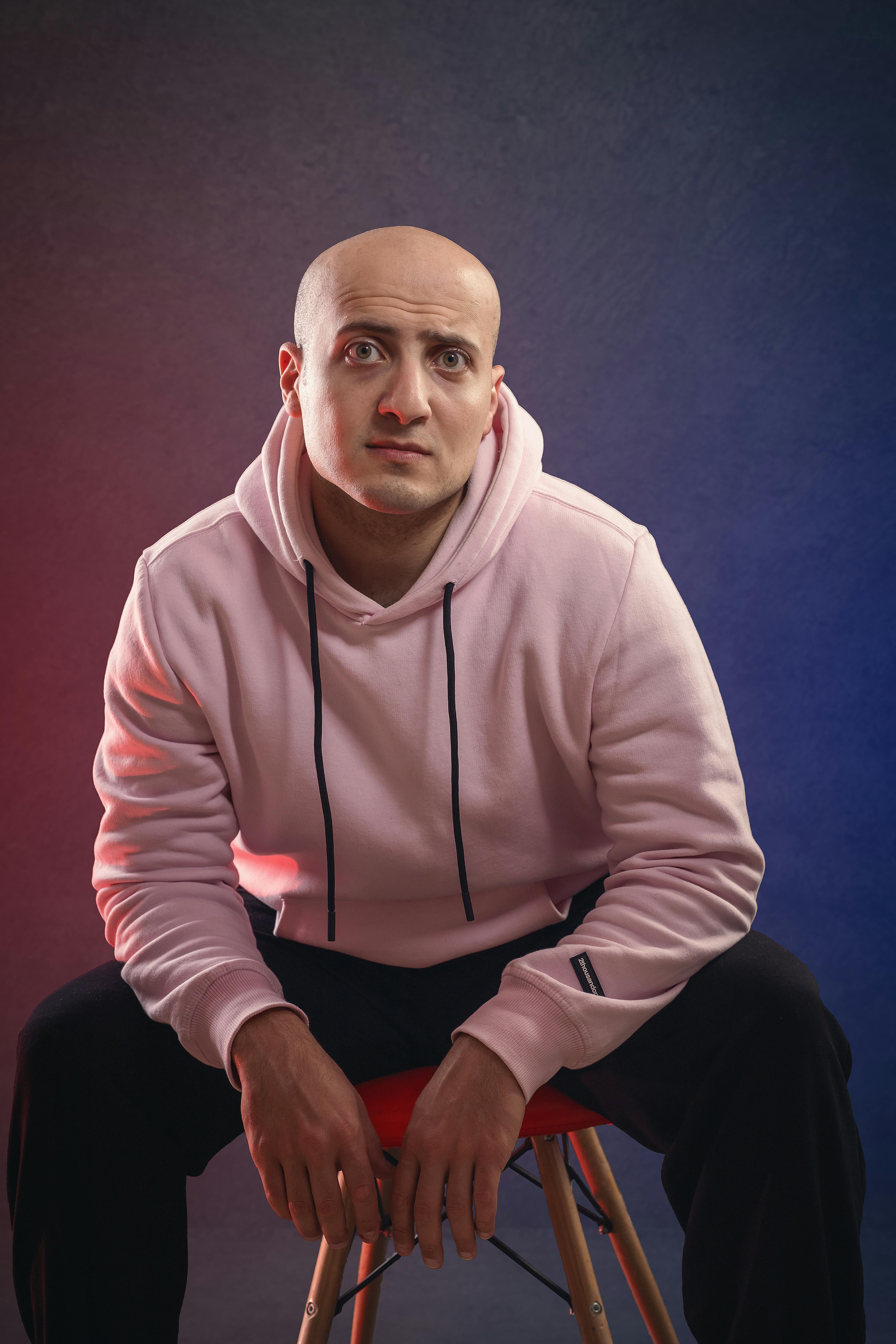 Portrait of a bald man wearing a pink hoodie seated on a stool in a studio setting.