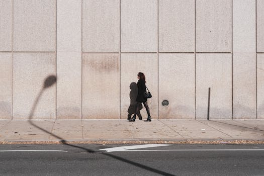 A woman in black walking past a concrete wall in an urban setting with shadows created by streetlights.