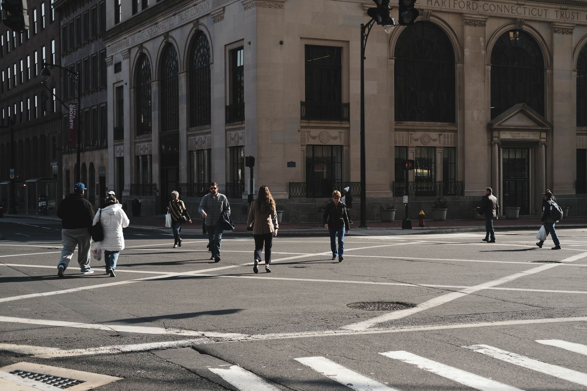 People crossing a city street with historical buildings in the background on a sunny day.