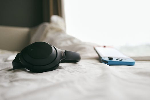 Close-up of a black headset and a gaming console resting on a soft, white bedspread in a calm, indoor setting.