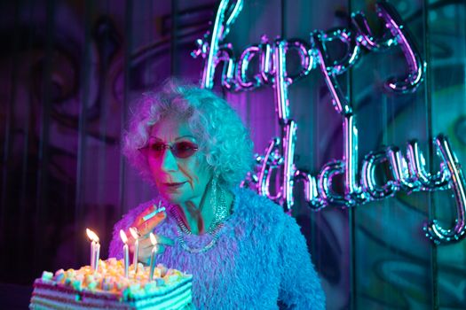 Elderly woman with grey hair celebrating birthday, surrounded by vibrant neon lights and birthday cake.