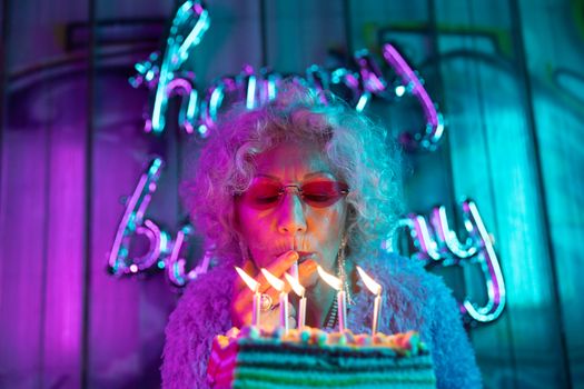 Trendy senior woman blowing out candles at a vibrant birthday celebration.