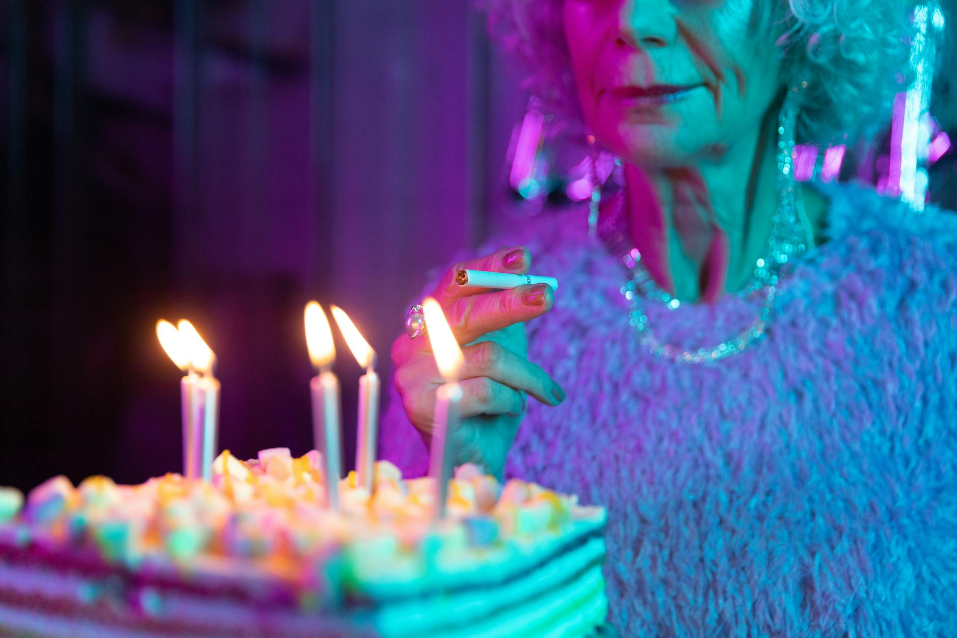 A senior woman holds a cigarette near a birthday cake with lit candles in a neon-lit room.