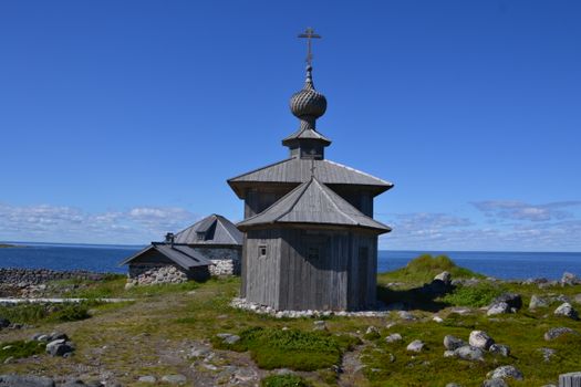 A rustic wooden chapel stands near the scenic Barents Sea under a bright blue sky.