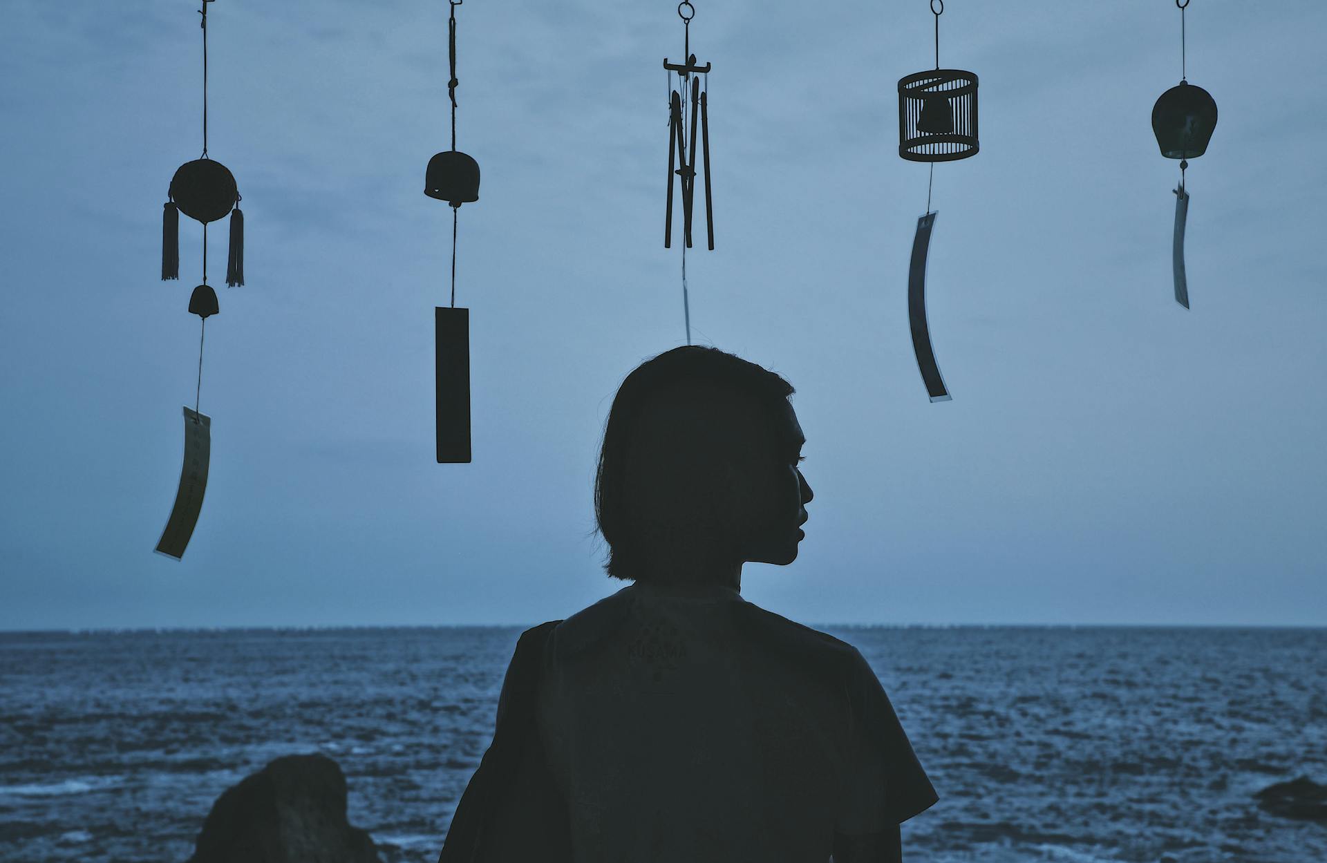 A silhouette of a young woman by the ocean with traditional Japanese wind chimes at dusk.