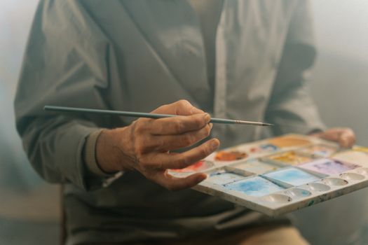 A close-up shot of an artist's hand holding a paintbrush and palette filled with vibrant watercolors.