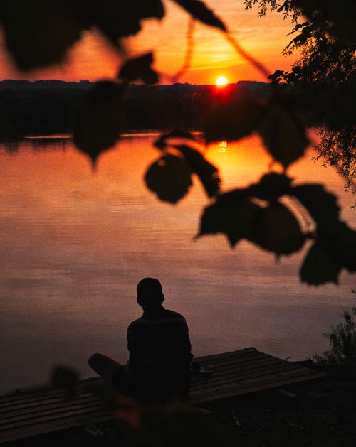 Free Silhouette of a person sitting by a peaceful lake during a vibrant sunset, creating a serene landscape. Stock Photo