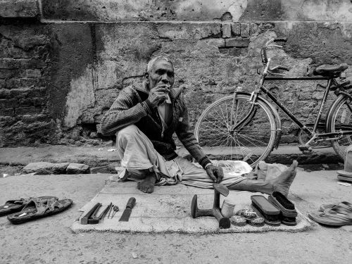 Free Black and white photo of an elderly cobbler sitting beside a bicycle, showcasing street life. Stock Photo