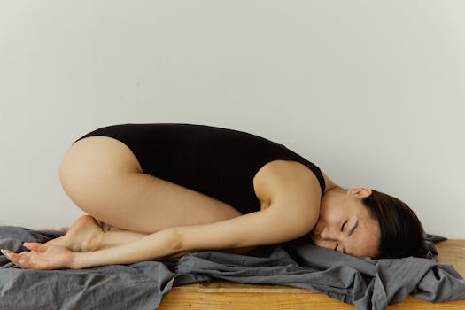 An Asian woman in a black bodysuit is resting peacefully on a gray blanket against a white wall.