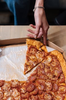 A hand reaches for a slice of delicious pepperoni pizza in a cardboard box.