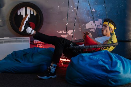Young man enjoys gaming on a console while relaxing in a hanging chair indoors.