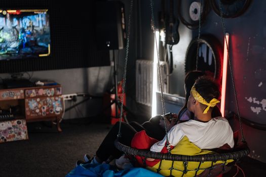Two friends gaming indoors on a comfy swing chair, enjoying leisure time.