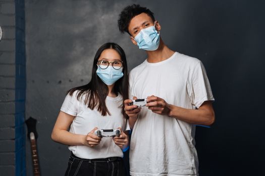 Two young adults playing video games indoors, wearing face masks for safety.