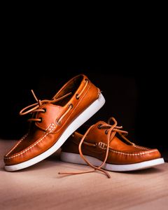 Close-up of brown leather boat shoes with white soles on a wooden floor against a black background.