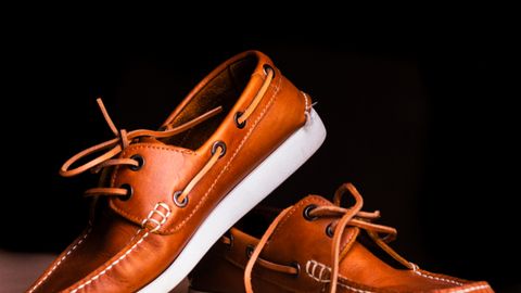 Close-up of brown leather boat shoes with white soles on a wooden floor against a black background.