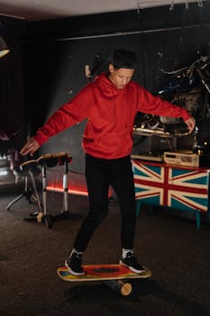 A young man in a red hoodie practices balance on a skateboard indoors.