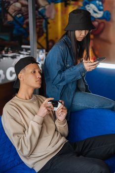 Two young adults playing video games and using a smartphone while relaxing indoors.