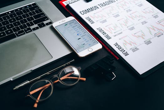 Workstation with laptop, smartphone, eyeglasses, financial charts, and pen for trading analysis.