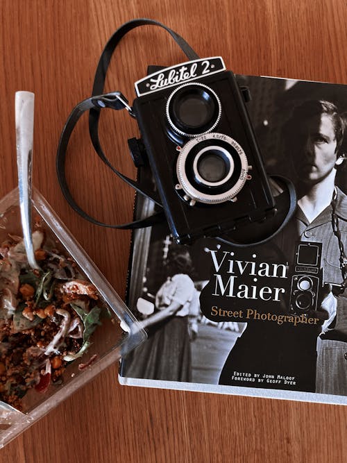 Top view of vintage photo camera and magazine placed on wooden table near plastic container with takeaway salad