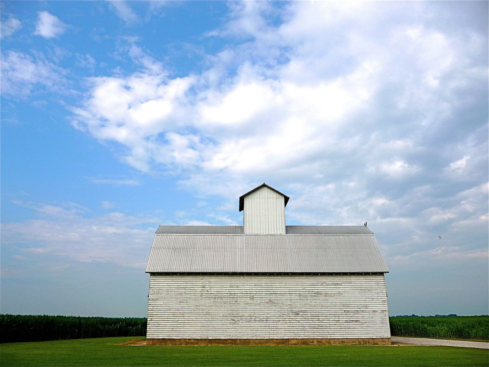 A rustic white barn stands against a backdrop of blue sky and clouds in a rural farmland setting.