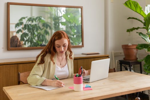 Red-haired woman writing at a desk with a laptop, in a bright modern office setting.