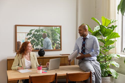 Man Talking to a Colleague while Sitting on Wooden Desk