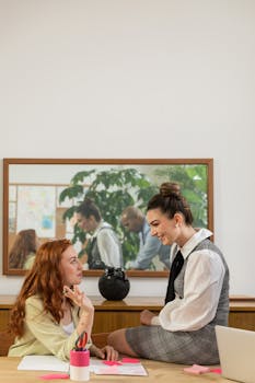 Two women having a discussion at a desk in an office setting with a reflection in the background.