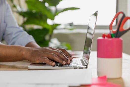Close-up of male hands typing on a laptop at a desk, surrounded by office supplies and plants.