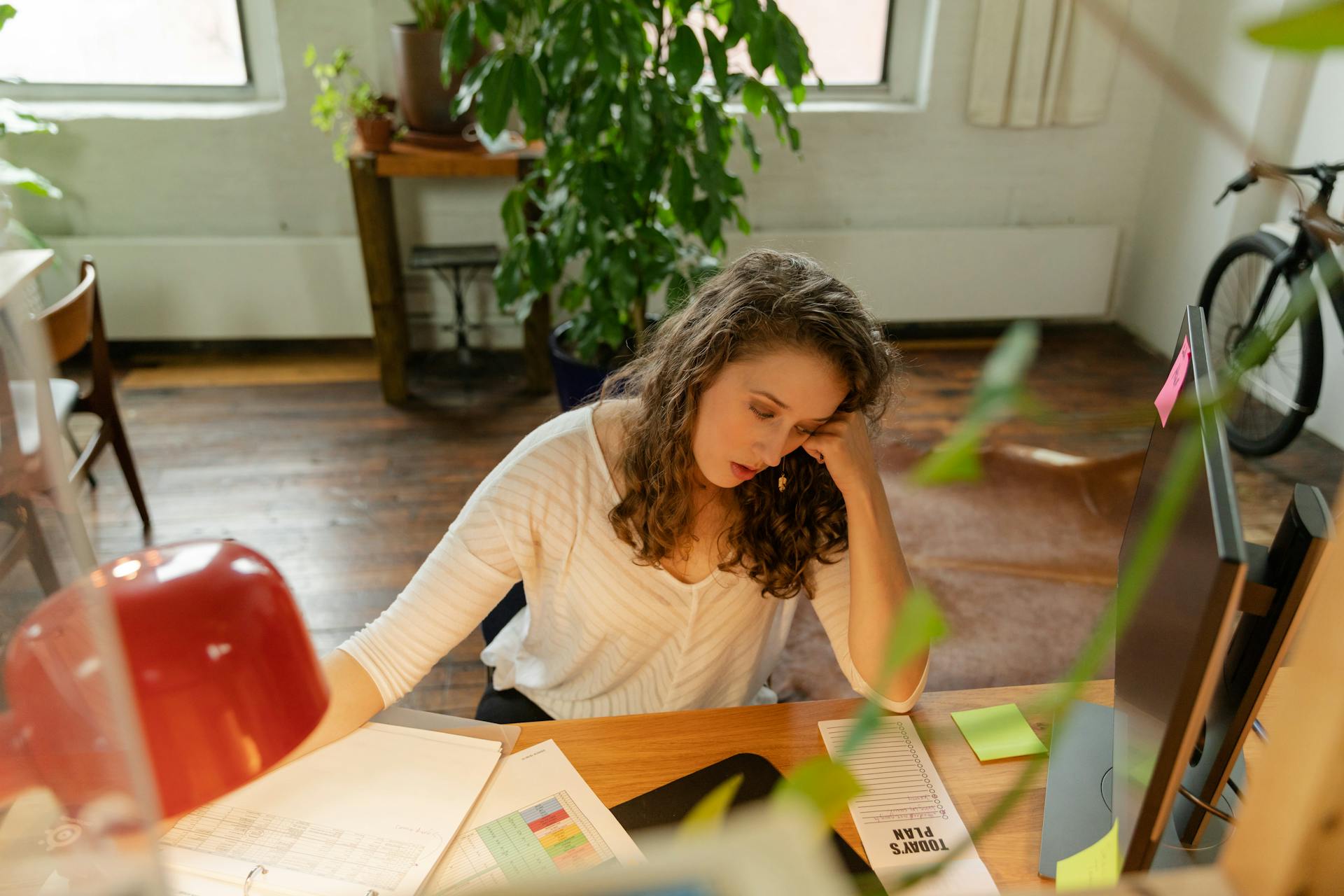 A woman sits at a wooden desk, appearing tired amid office work with sticky notes and a monitor.